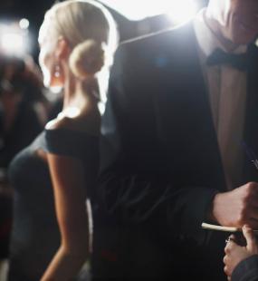 A man in a tuxedo signs an autograph book while a blonde woman in a ballgown is photographed by paparazzi behind him