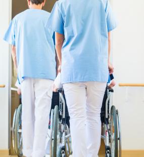 Two care workers in nurse scrubs push two wheelchairs through the corridor of a care home